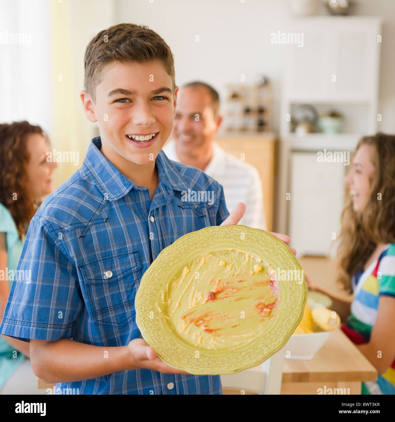 Ragazzo sorridente visualizzando la piastra a vuoto con la famiglia a tavola in background Foto Stock