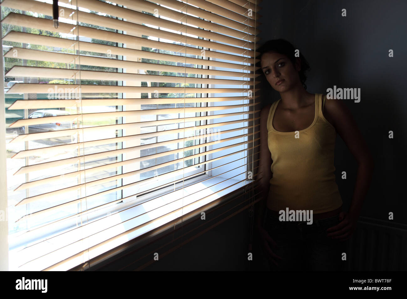 Giovane donna in piedi da solo in una stanza buia con una tenda a veneziana, guardando verso la telecamera. Foto Stock