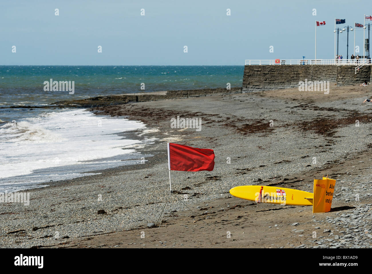 Red Flag di avviso con RNLI Salvataggio tavola da surf su una spiaggia deserta con mare mosso, Aberystwyth, Galles Foto Stock