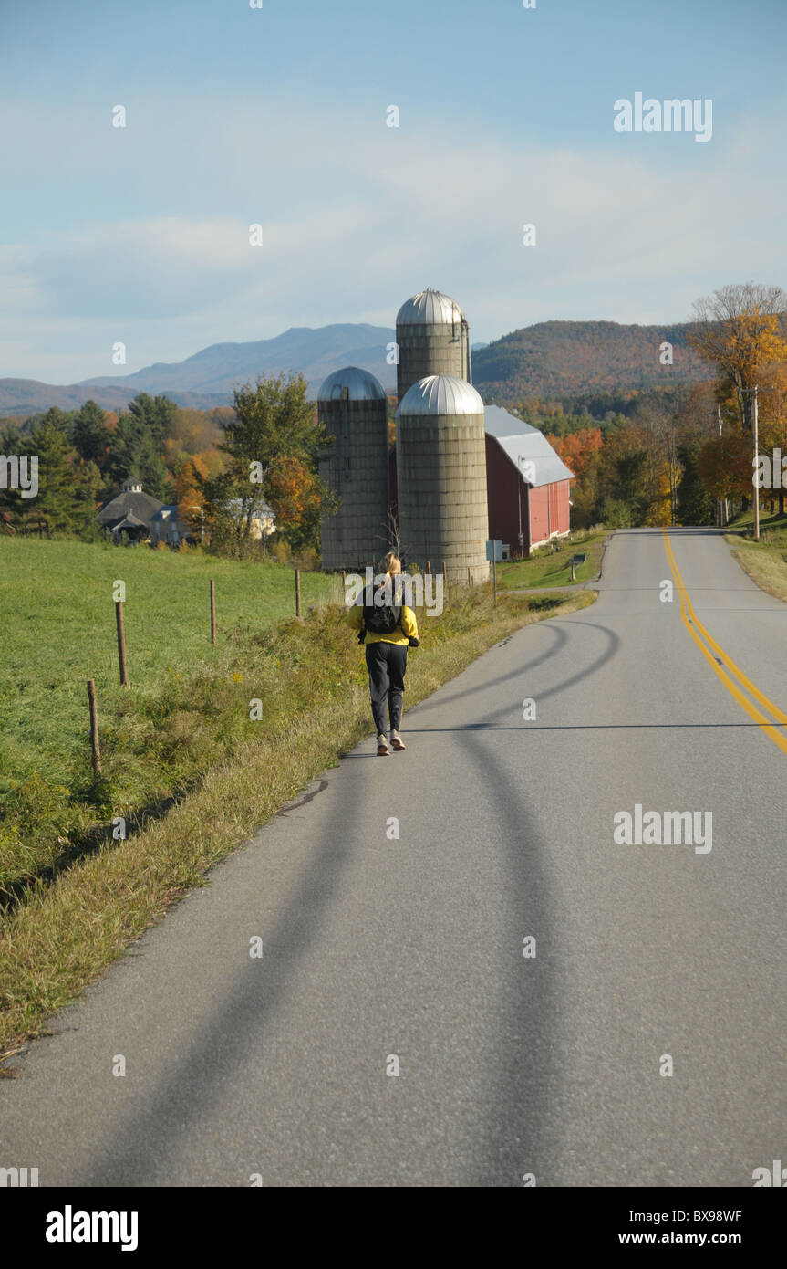 Una donna corre lungo una strada di campagna con una fattoria in background in Waitsfield, Vermont Foto Stock