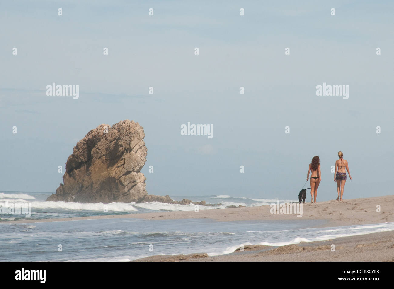 2 distante figure in abiti nuotare per passeggiare sulla spiaggia di sabbia in un giorno nuvoloso con un cane legato passando una pila di roccia Foto Stock