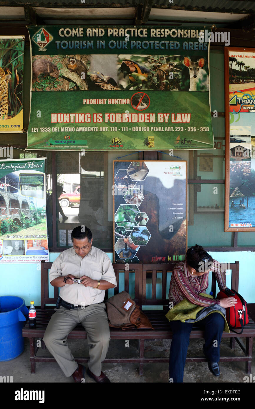I passeggeri in attesa in aeroporto sotto segno in inglese incoraggiando il turismo responsabile e dichiarando la caccia è proibita, Rurrenabaque, Bolivia Foto Stock