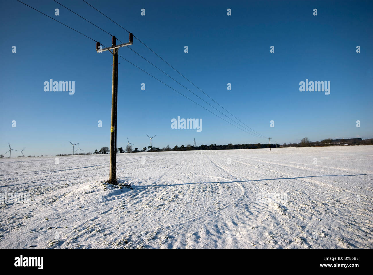 Singolo palo del telegrafo in piedi in una coperta di neve campo con un profondo cielo blu, in Inghilterra. Le turbine eoliche lungo l'orizzonte. Foto Stock