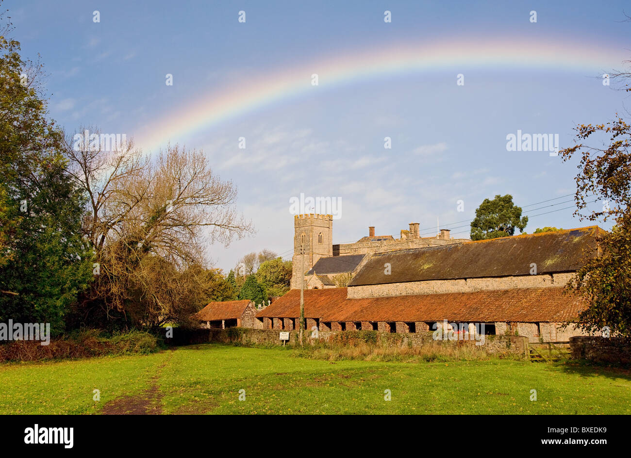 Rainbow oltre la chiesa di St Mary in East Quantoxhead nel Somerset con edifici agricoli confinante villaggio verde Foto Stock