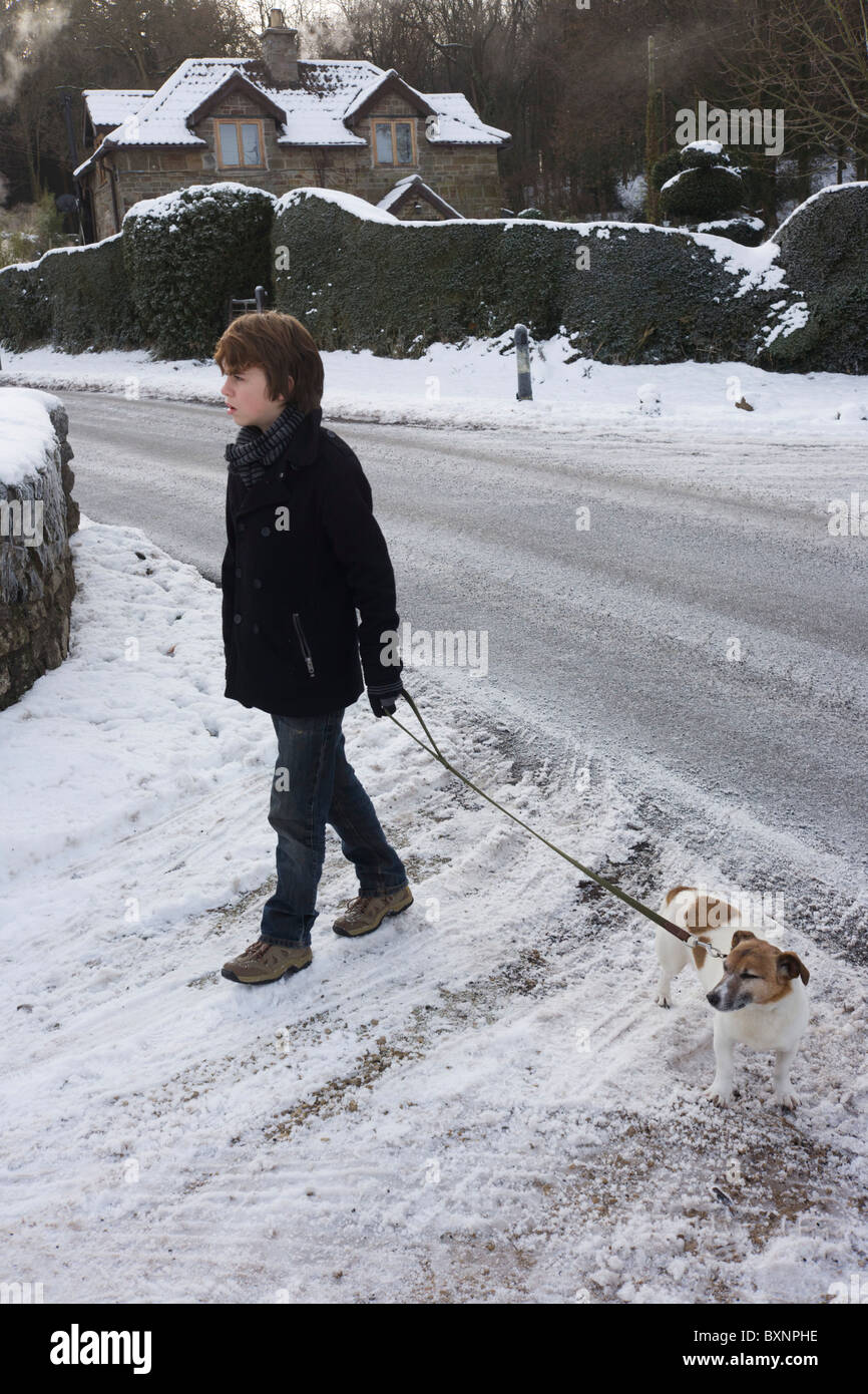 Giovani 12 anno-vecchio ragazzo circa a croce strada ghiacciata a camminare un pet Terrier cane durante inverno nevica. Foto Stock