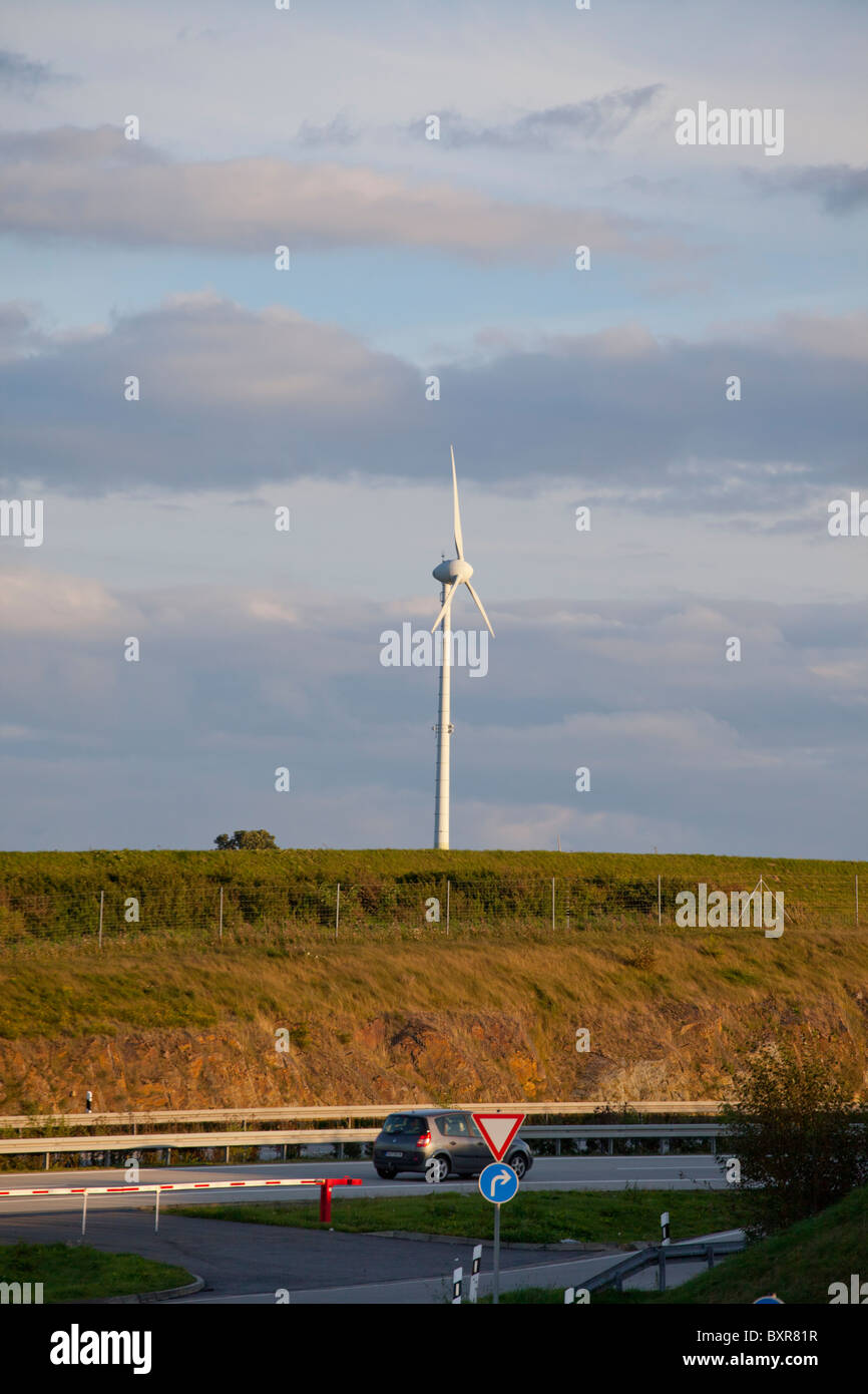 Turbina eolica in piedi su una collina erbosa vicino a una strada con un'auto in Germania sotto un cielo nuvoloso. Foto Stock