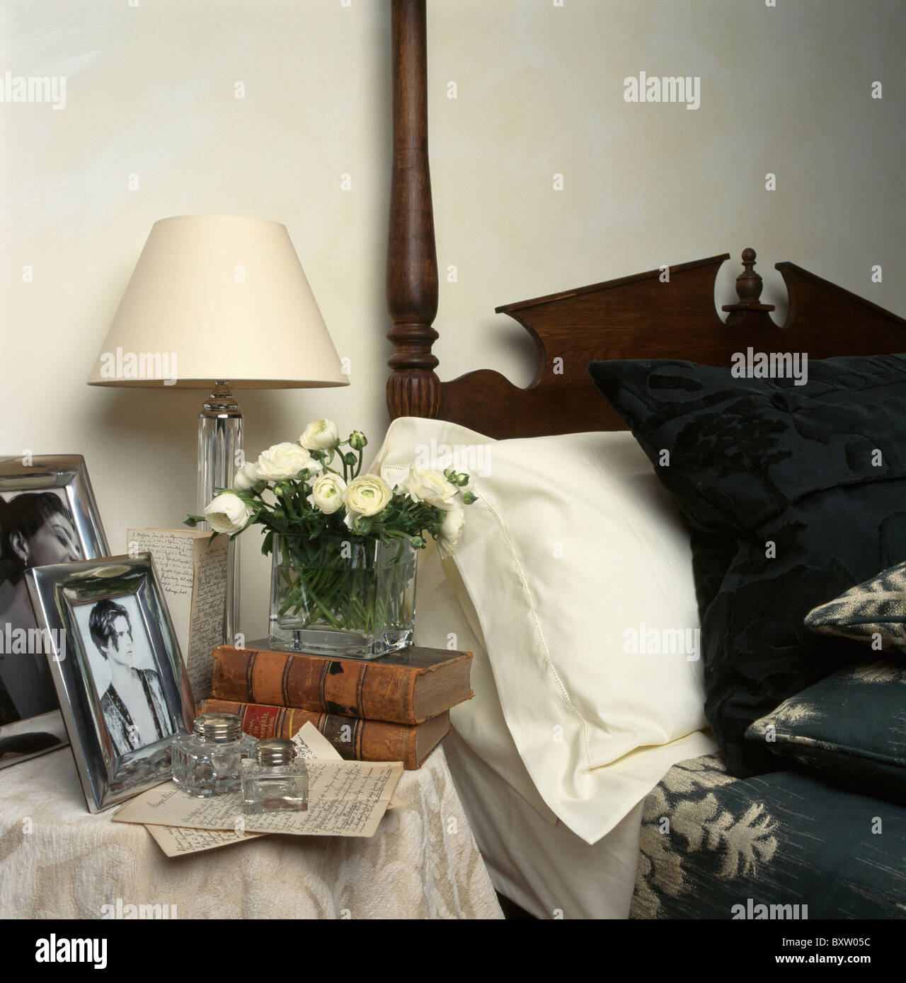 Close-up di crema lampada e fotografie incorniciate con libri sul comodino accanto al letto con cuscino bianco e modellato bed-cover Foto Stock