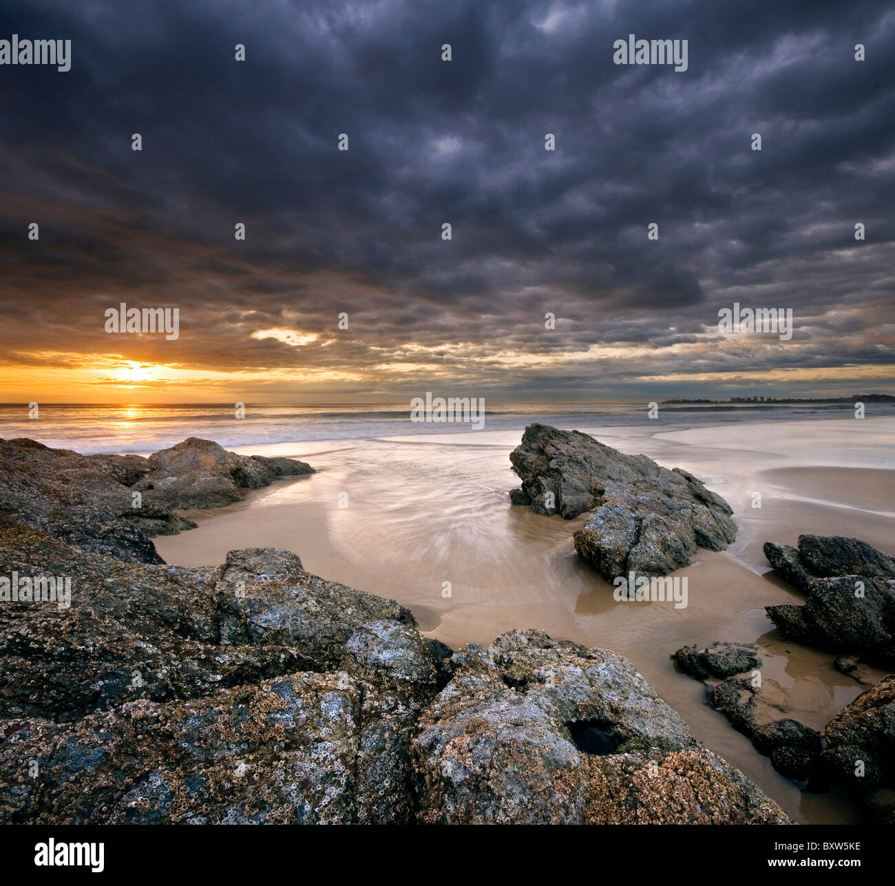 Rocce sulla spiaggia al tramonto con cielo drammatico sul formato quadrato Foto Stock