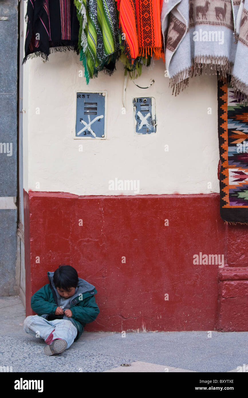 Lonely bambino seduto a terra contro la parete di un locale negozio di artigianato di Puno, Perù Foto Stock