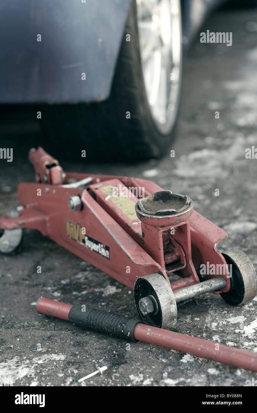 Una chiusura di un idraulico rosso cric idraulico a carrello accanto a un auto su un gelido viale con il manico staccata. Foto Stock