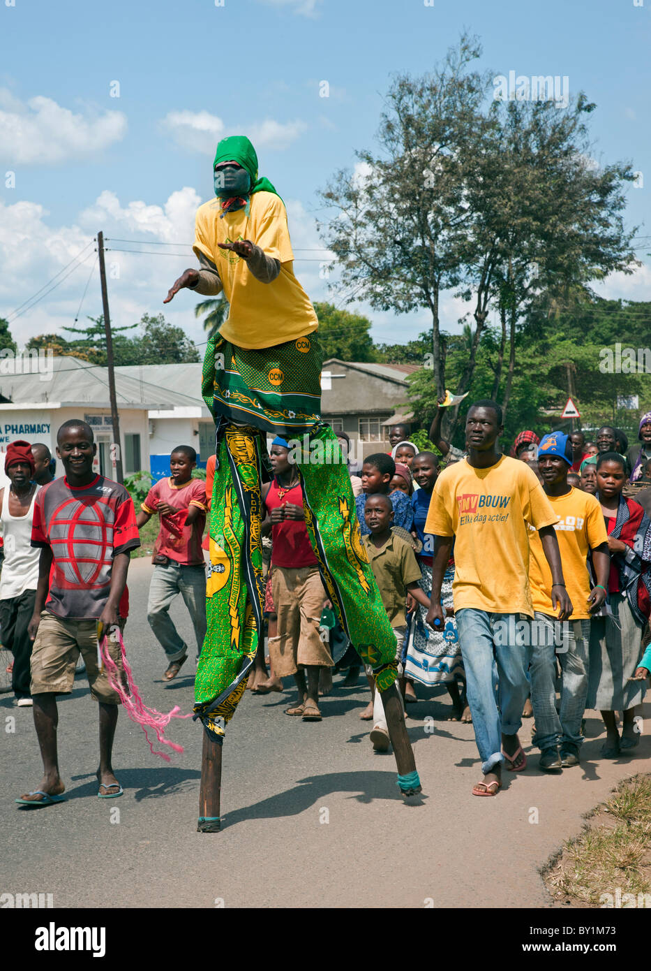 Un stilt-walker è a capo di un gruppo di giovani e bambini per le strade di Kerogwe prima di una politica dei rally. Foto Stock