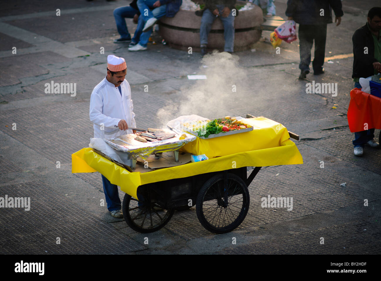 Balik Ekmek Fish Sandwiches Street Vendor Istanbul Turkey // ISTANBUL, Turkey - Un venditore ambulante grigia il pesce fresco su un carrello mobile al mercato del pesce di Karakoy vicino al ponte Galata a Istanbul. Questi venditori preparano tipicamente balık ekmek (panini di pesce tradizionali), un popolare cibo di strada preparato con pesce appena pescato dalle acque locali. La pratica di grigliare il pesce lungo il lungomare rappresenta una tradizione culinaria di lunga data a Istanbul. Foto Stock
