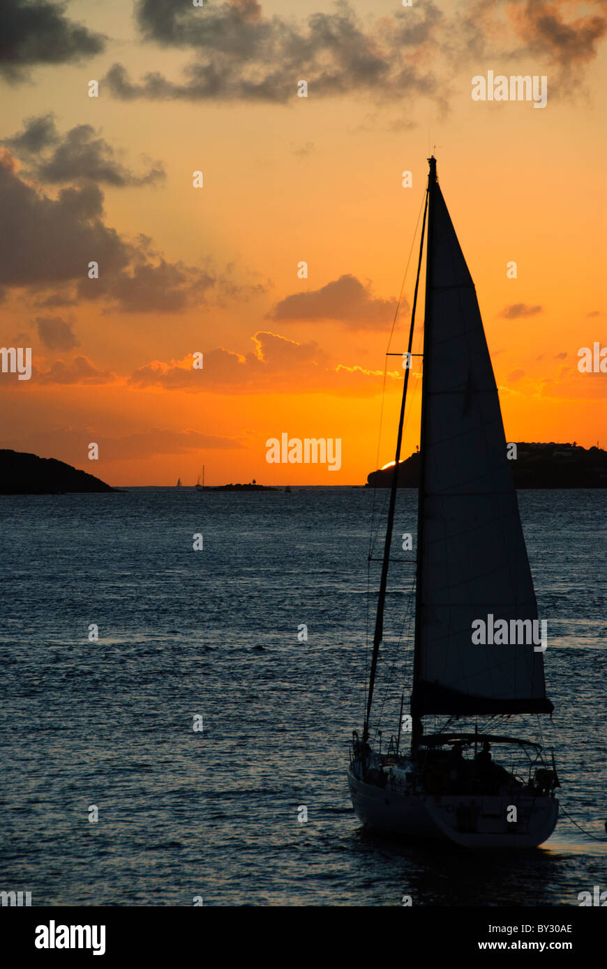 ISOLE VERGINI AMERICANE: Una barca a vela solitaria scivola pacificamente attraverso le acque calme verso uno spettacolare tramonto caraibico nelle Isole Vergini americane. La sagoma della barca è drammaticamente contrastata con le vivaci arance, i rosa e i viola che dipingono il cielo serale. Foto Stock