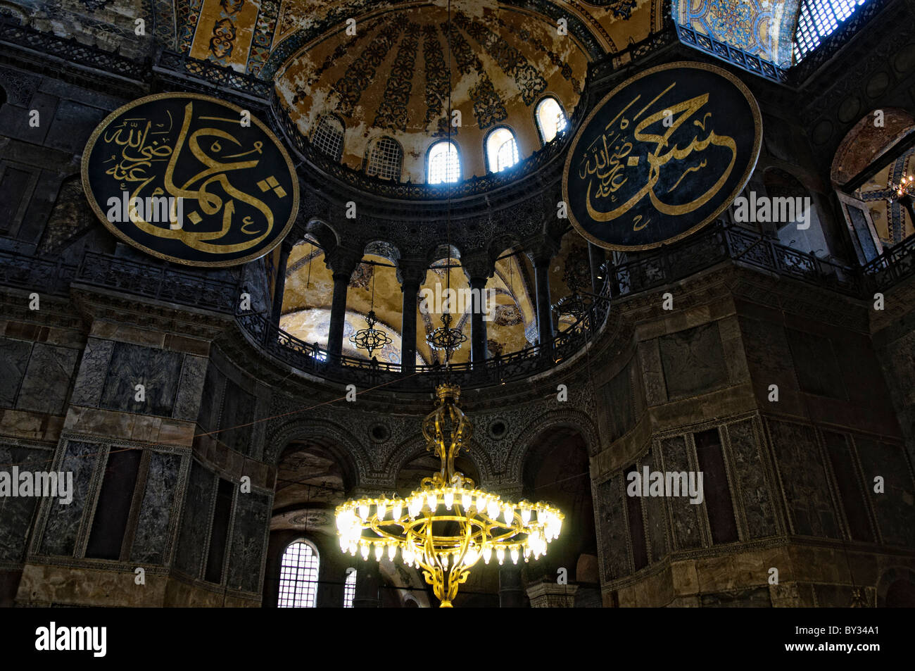 ISTANBUL, Turchia: gli ornati avize (lampadari turchi) sono appesi al soffitto a cupola della Basilica di Santa Sofia, illuminando il vasto spazio interno. Questi apparecchi di illuminazione in bronzo di epoca ottomana sono stati progettati per fornire illuminazione per i servizi di preghiera. L'imponente cupola si innalza a 180 metri dal pavimento, con l'avize sospeso a varie altezze per creare strati di luce. Foto Stock