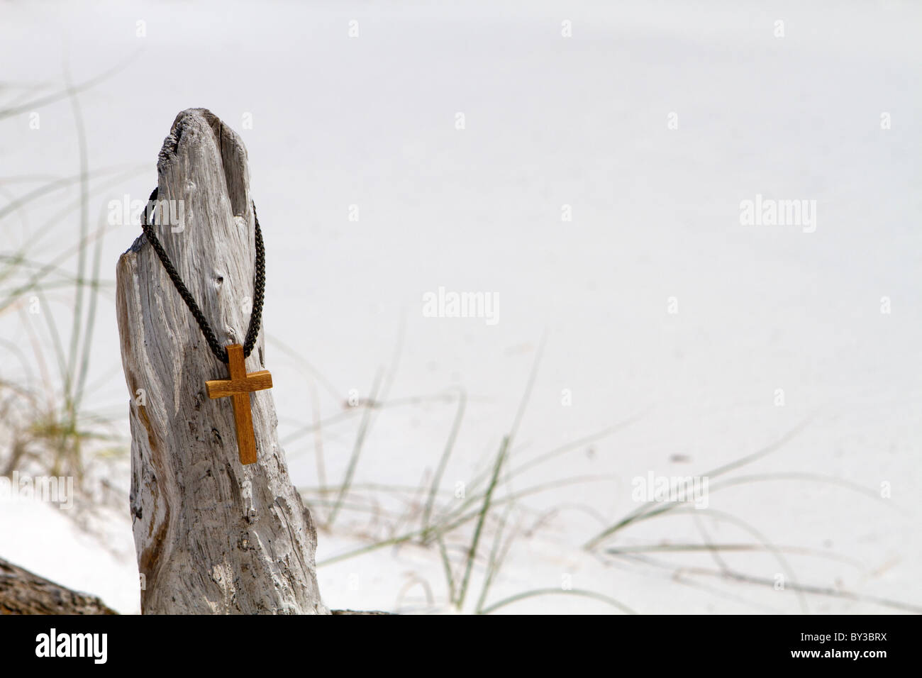 Croce pende da una corda intrecciata collana su un pezzo di driftwood con una spiaggia di sabbia sullo sfondo. Foto Stock