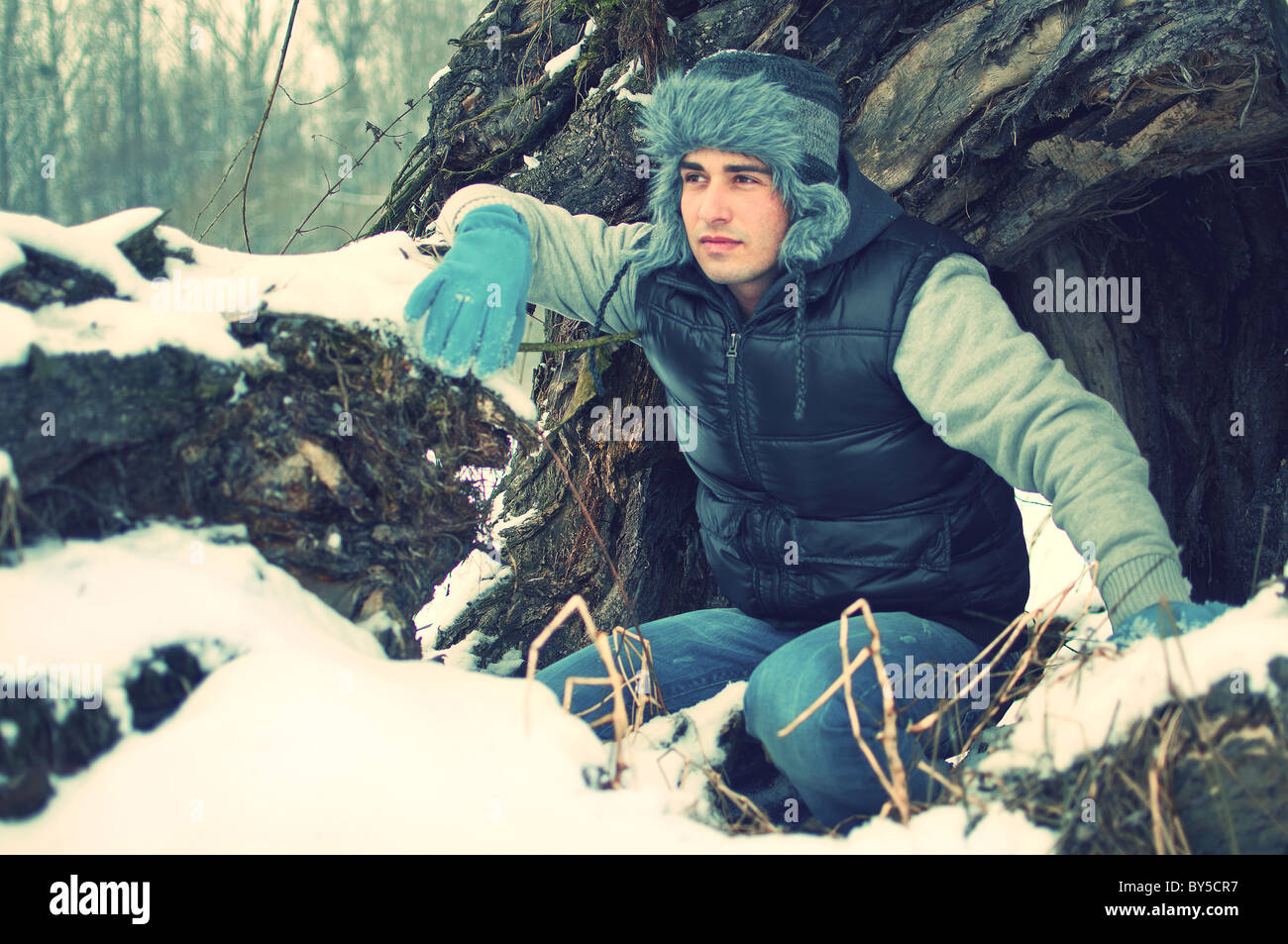 Ritratto di moda di un accovacciato bel giovane guardando qualcosa in inverno davanti al grande vecchio tronco di albero Foto Stock