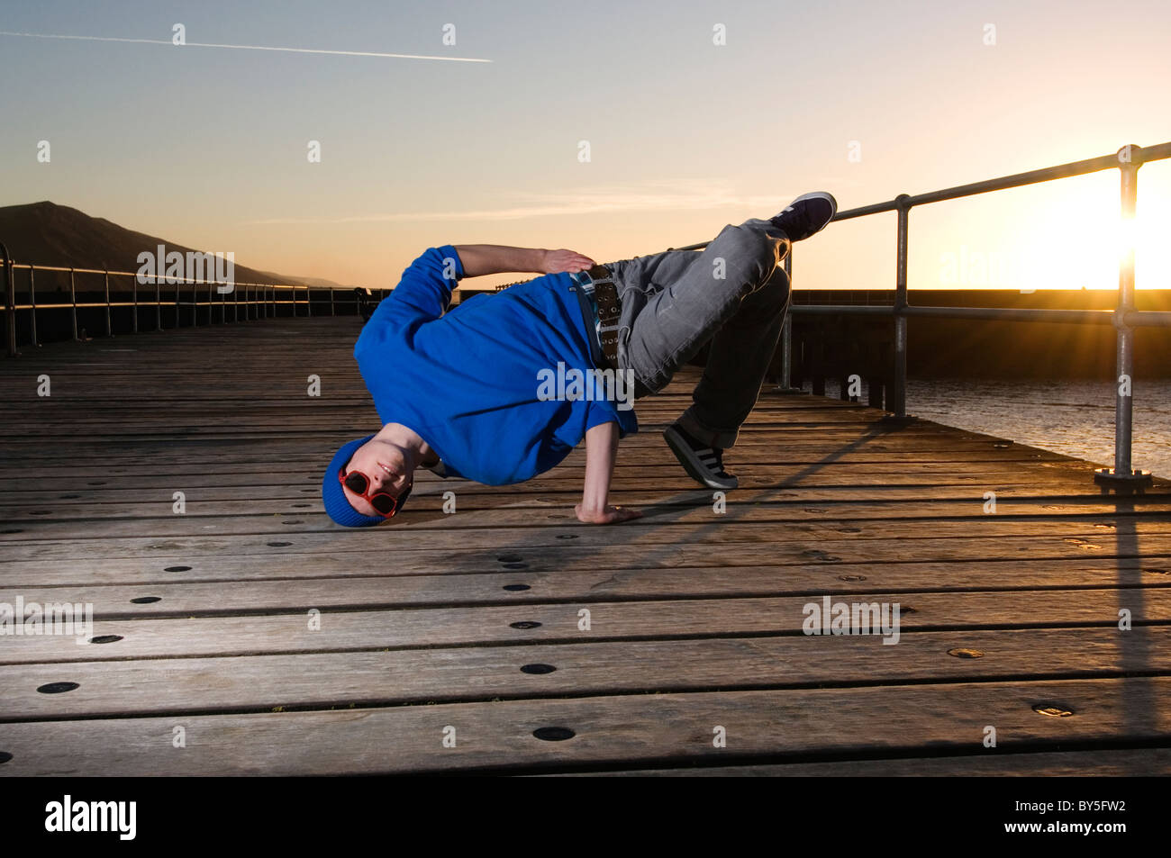 Un ampio angolo di inquadratura di un giovane uomo di eseguire la sedia "Freeze" sull'un molo mentre il sole tramonta. Foto Stock