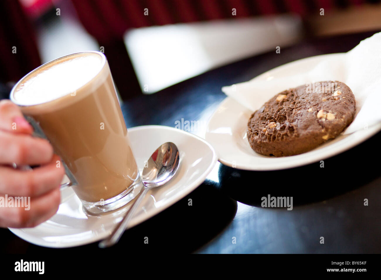 Una donna di mano afferra un caffelatte bevanda caffè accanto a un cookie al cioccolato biscotto. Foto Stock Una donna di mano afferra un caffelatte bevanda caffè accanto a un cookie al cioccolato biscotto. Foto Stock