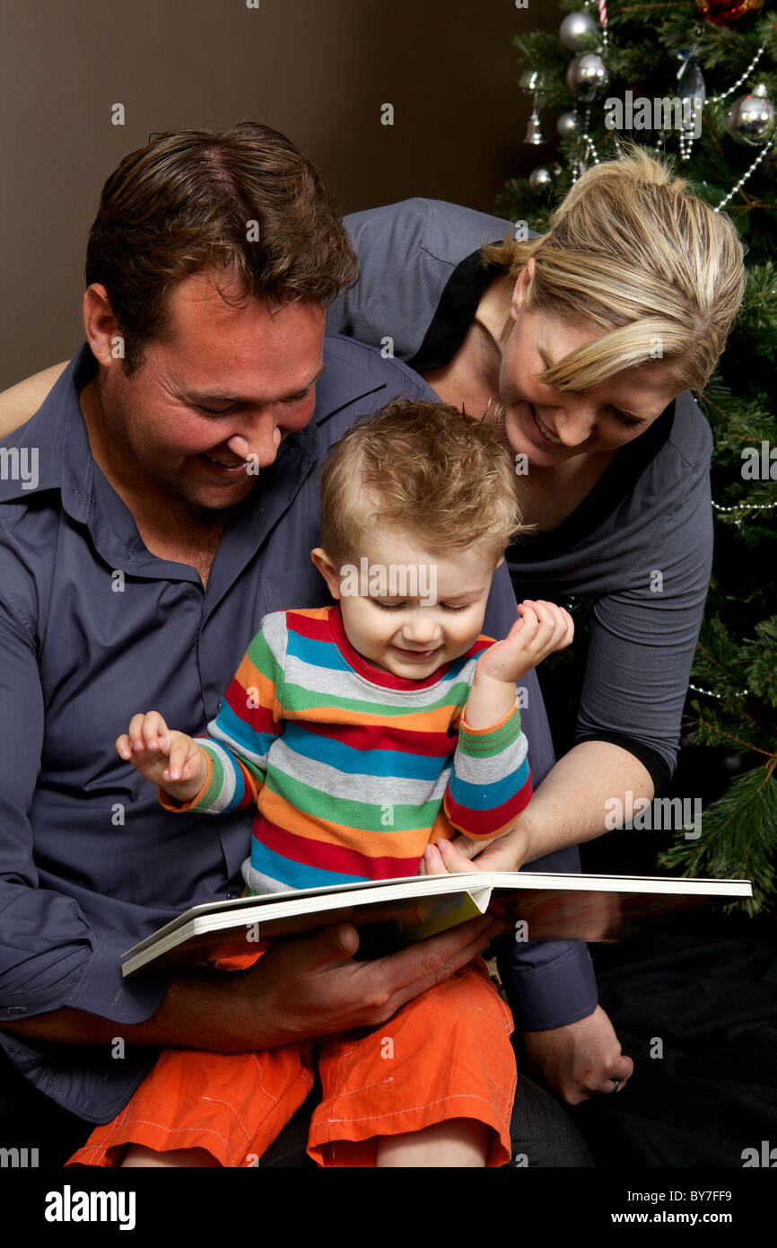 Il padre e la madre la lettura di un libro insieme con il loro bambino figlio al tempo di Natale con l'albero di Natale Foto Stock