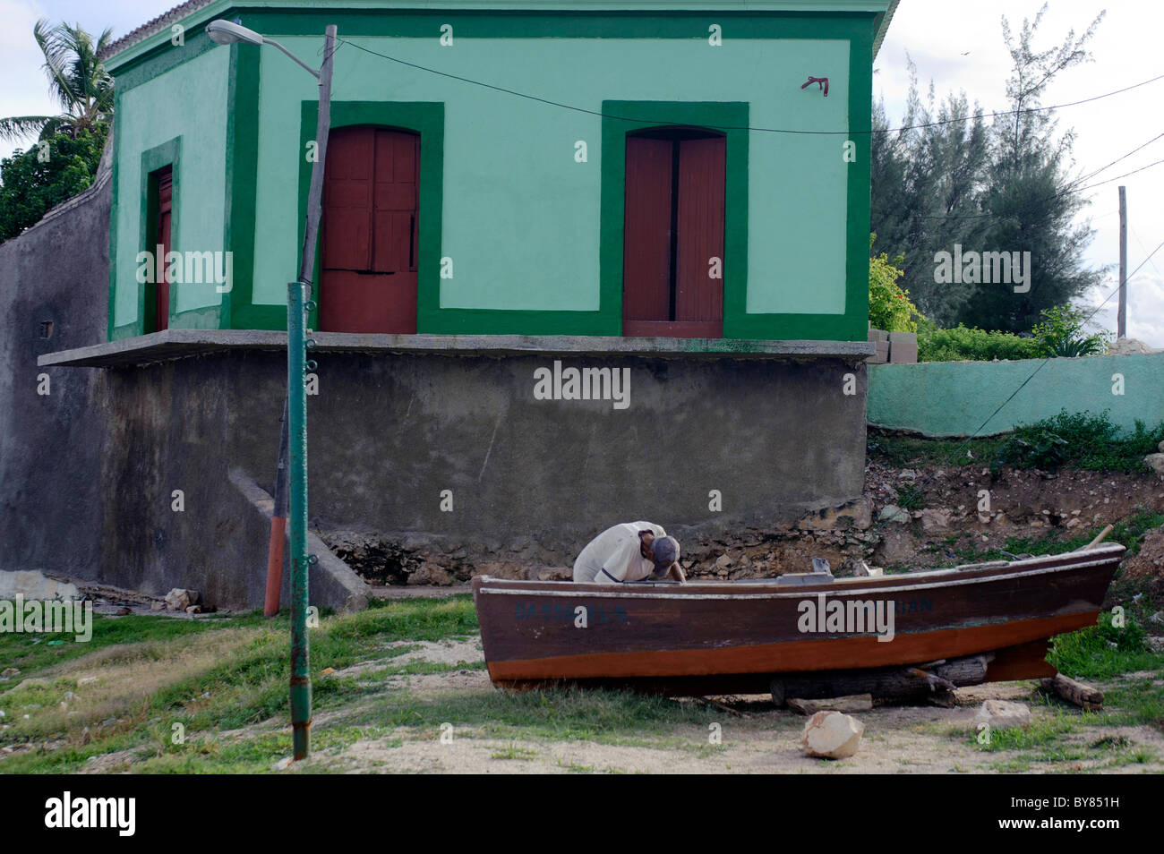 Vecchio Pescatore cubano con t-shirt e cappuccio a Cuba la pittura la sua barca. Casa dipinta in background Foto Stock