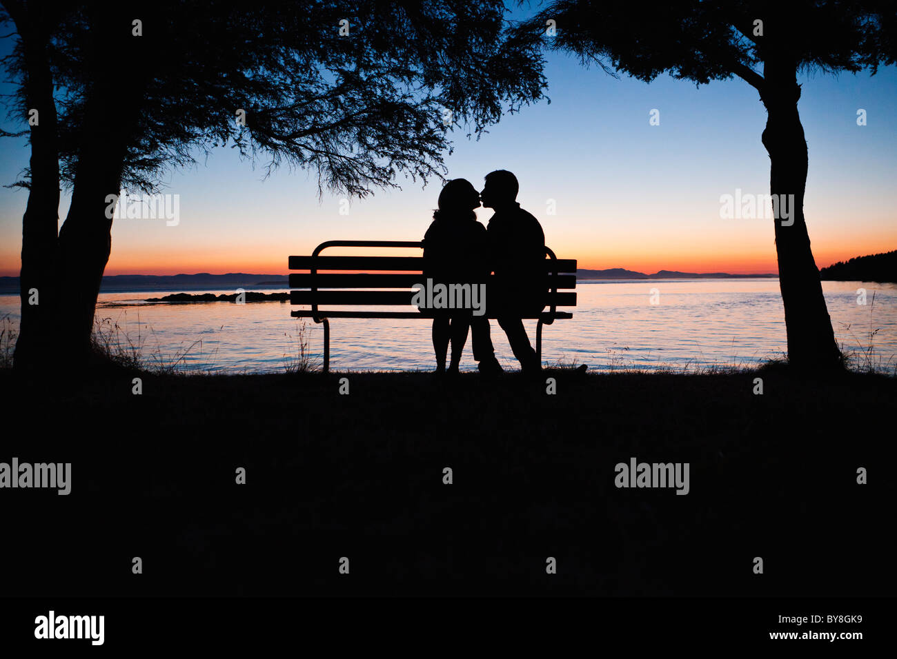 Un uomo e una donna baciare su una panchina nel parco al tramonto. Parcheggio contea di San Juan Island, Washington, Stati Uniti d'America. Foto Stock
