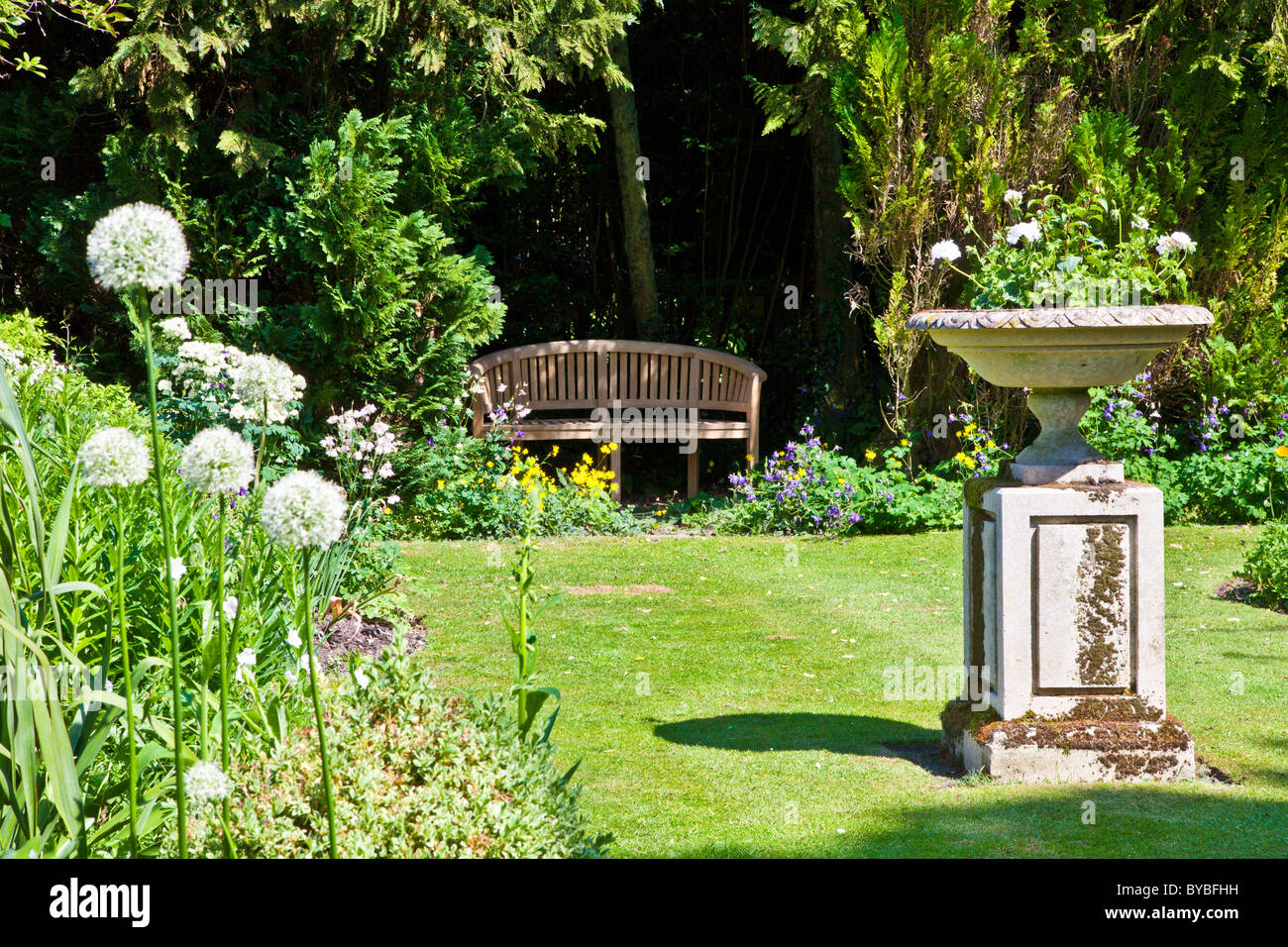 Un piccolo prato in un paese di lingua inglese giardino in estate con una panca in legno nell'ombra di una siepe e una piantatrice di pietra su un plinto. Foto Stock