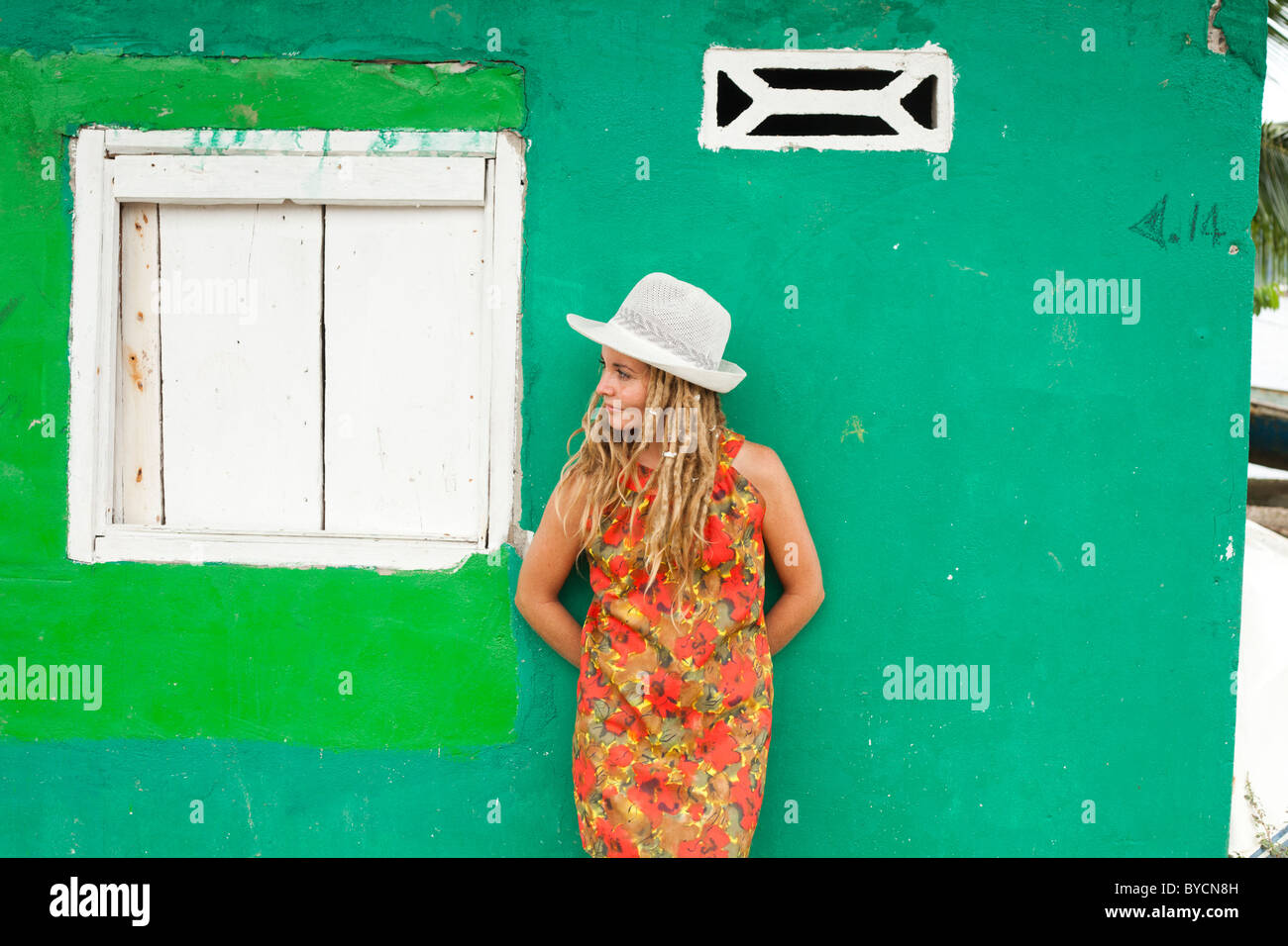 Donna bionda con dreadlocks in abito rosso e cappello bianco appoggiato contro una parete verde in Panama Foto Stock