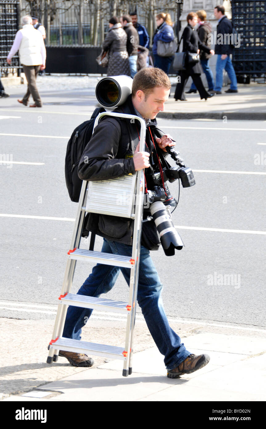 Fotografo maschio che trasportano fotocamere e lenti di attrezzature con scaletta camminando sul marciapiede case esterno del Parlamento Londra Inghilterra REGNO UNITO Foto Stock