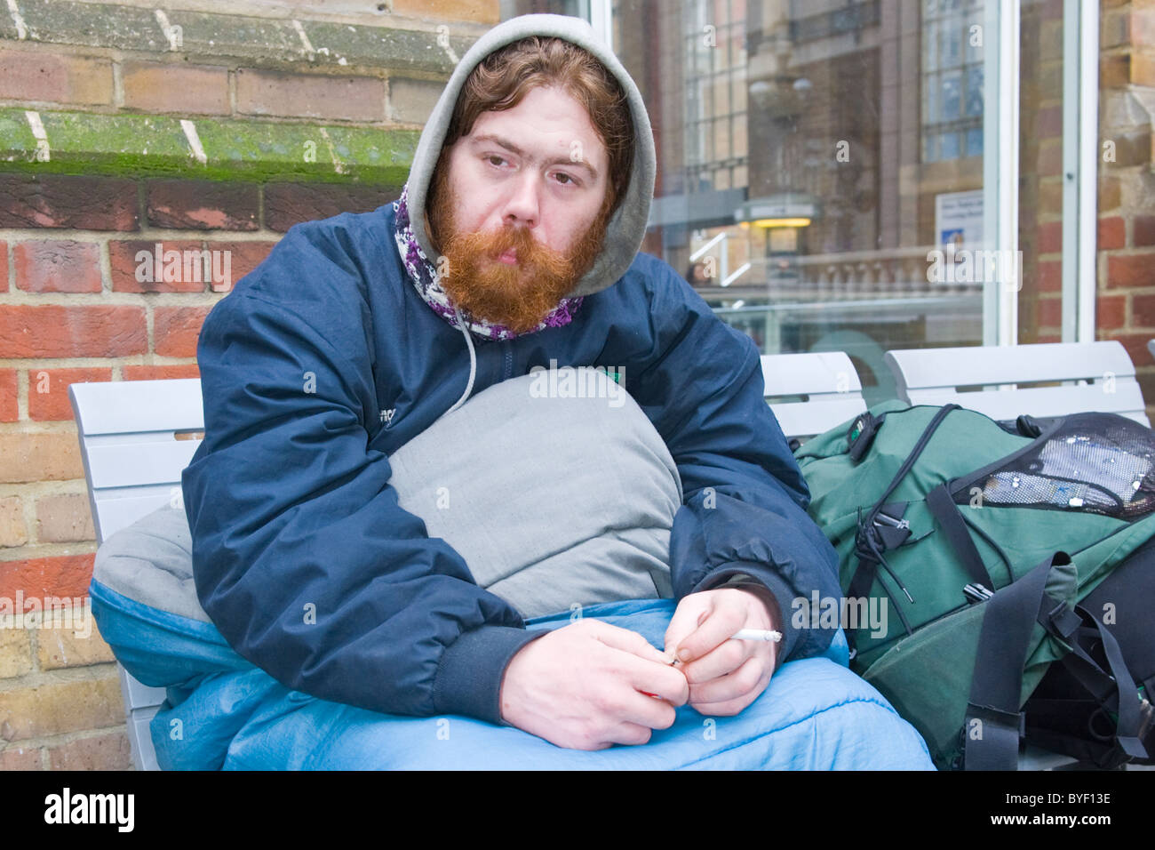 , Londra Liverpool Street Station , rosso intitolata & barbuto giovani senzatetto uomo avvolto nel sacco a pelo fumatori - ventole di 2 anni Foto Stock