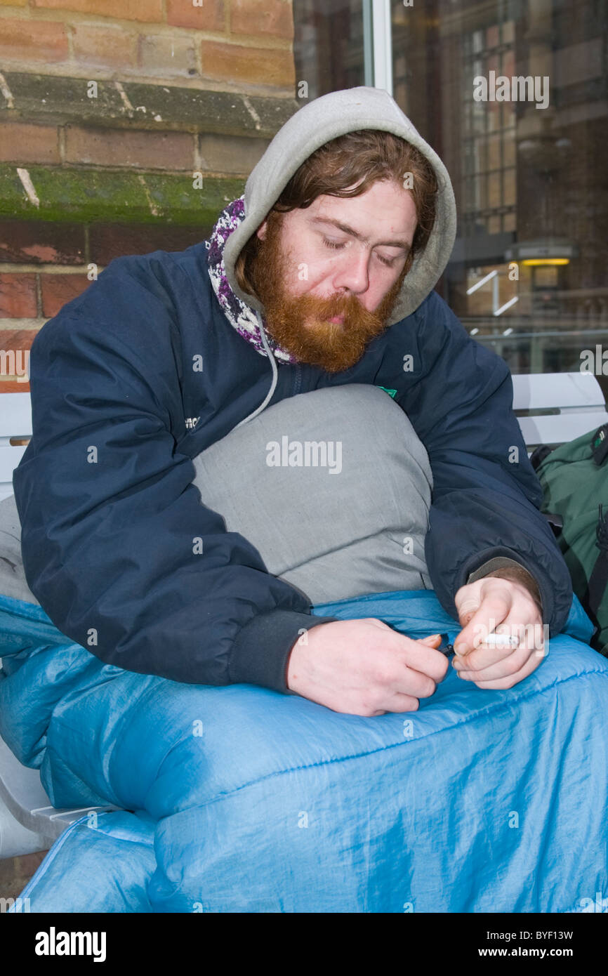 , Londra Liverpool Street Station , rosso intitolata & barbuto giovani senzatetto uomo avvolto nel sacco a pelo fumatori - ventole di 2 anni Foto Stock