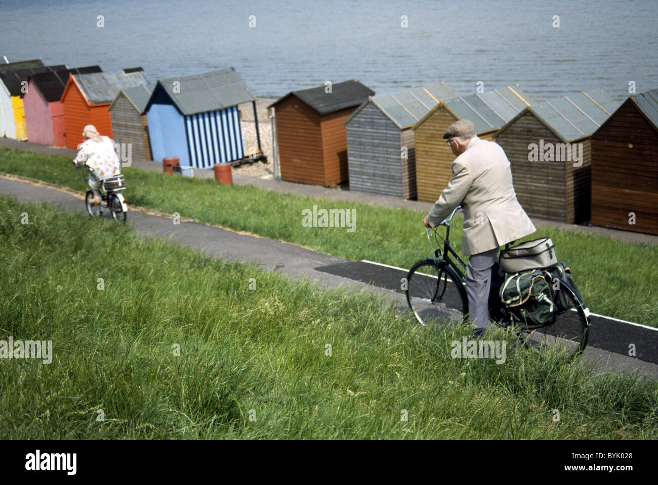 Due persone anziane in bicicletta lungo la riva del mare che si affaccia su Beach capanne Foto Stock