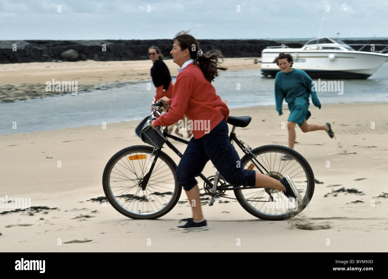 Spiaggia in scena con la donna in esecuzione con la sua mountain bike e due persone seguono in background Foto Stock