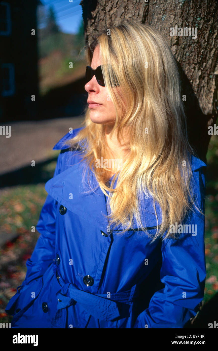 Giovane donna bionda in Blue Coat indossando occhiali da sole, appoggiata contro un albero, caduta, New Jersey Foto Stock