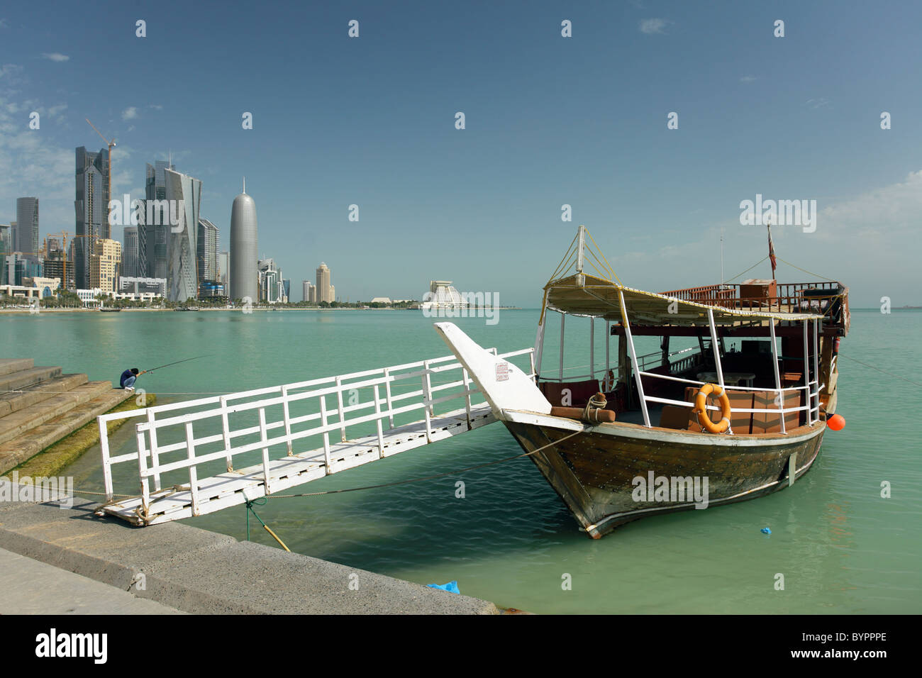 Un dhow attende su Doha Corniche, Qatar, per i turisti che desiderano un viaggio intorno alla baia, mentre un pescatore tenta la fortuna dalla riva. Foto Stock