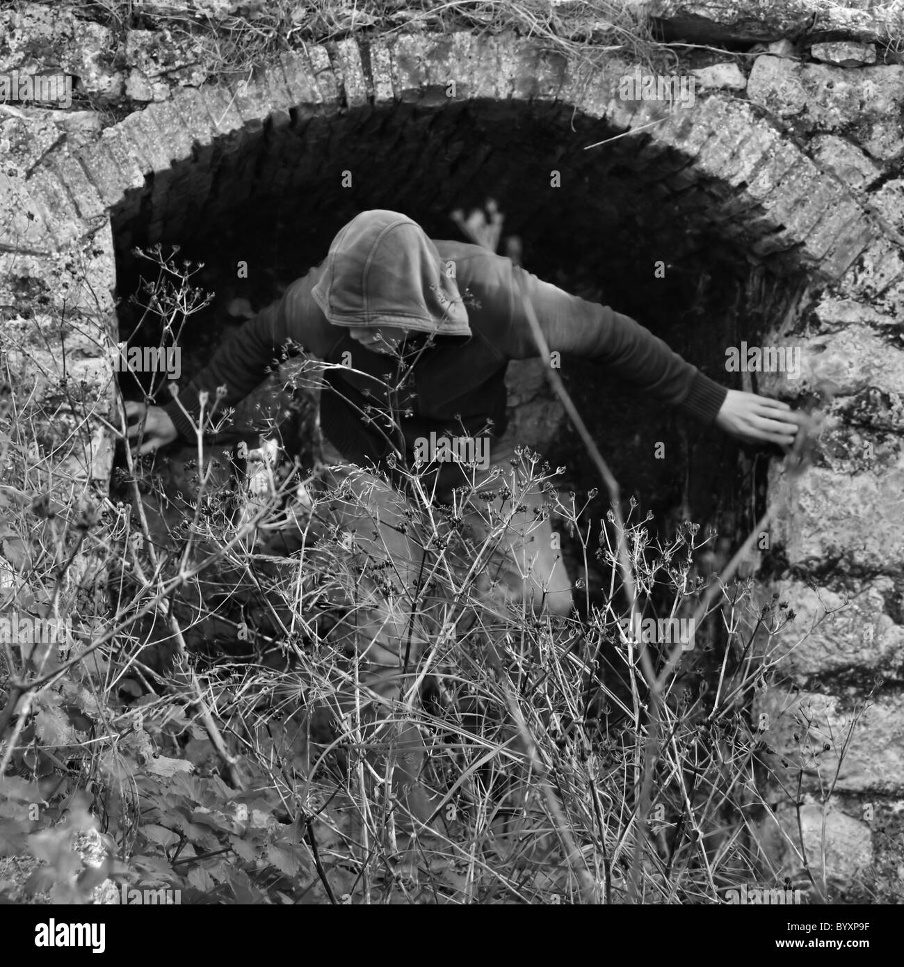 Uomo incappucciato oscurato da incolto piante sotto la cavità arcuata rurale della rovina. Foto Stock