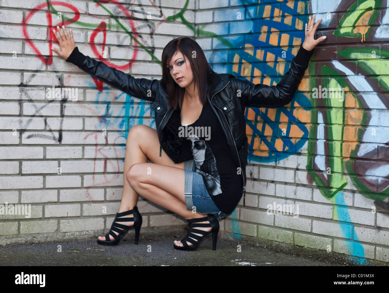 Giovane donna con i capelli scuri che indossa pantaloni a caldo, un nero  giacca di pelle e tacchi alti e in posa davanti un muro con graffiti Foto  stock - Alamy