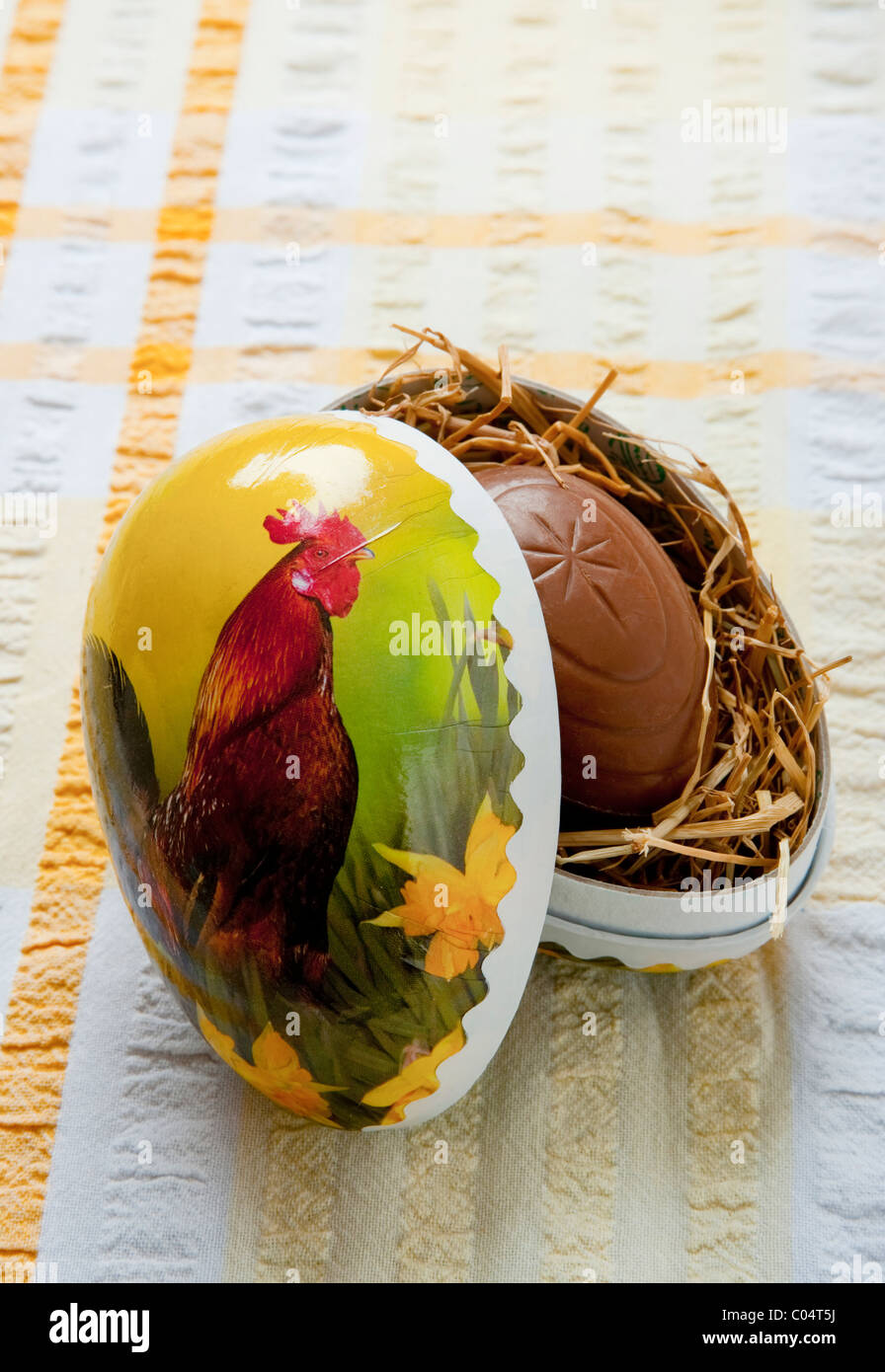 Francese cartone decorativo uovo di pasqua con uovo di cioccolato all'interno sul nido di fieno seduti sul giallo e tovaglia bianca Foto Stock
