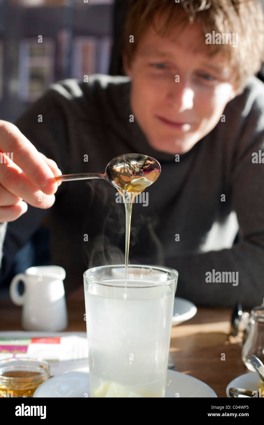 Miele e tè allo zenzero. Giovane uomo versando il miele in un bicchiere di succo di limone e il tè allo zenzero in una soleggiata cafe in Germania. Foto Stock