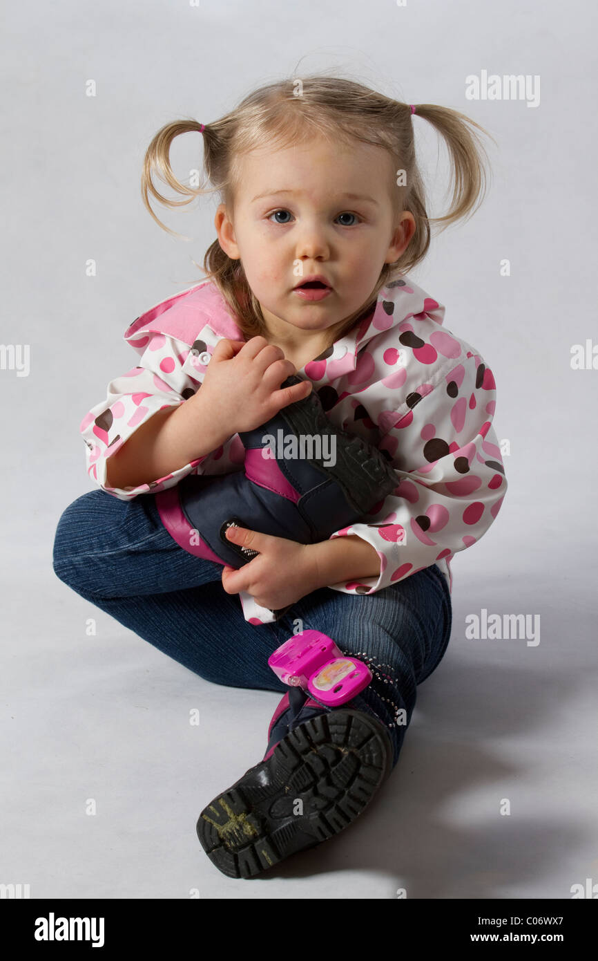 Due anni di bambino biondo con pig-tail sat holding il suo piede per il suo torace con lo sguardo verso la fotocamera con un giocattolo telefono rosa bilanciato su Foto Stock