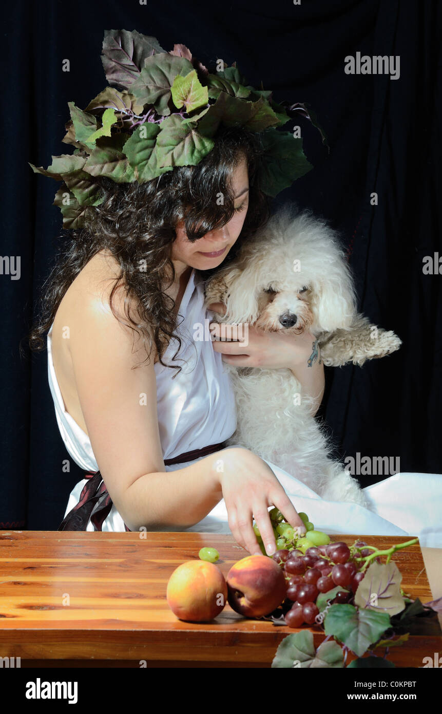 Donna attraente vestita come una femmina di Dionisio, il dio greco della vendemmia con la sua Bichon Frise. Foto Stock