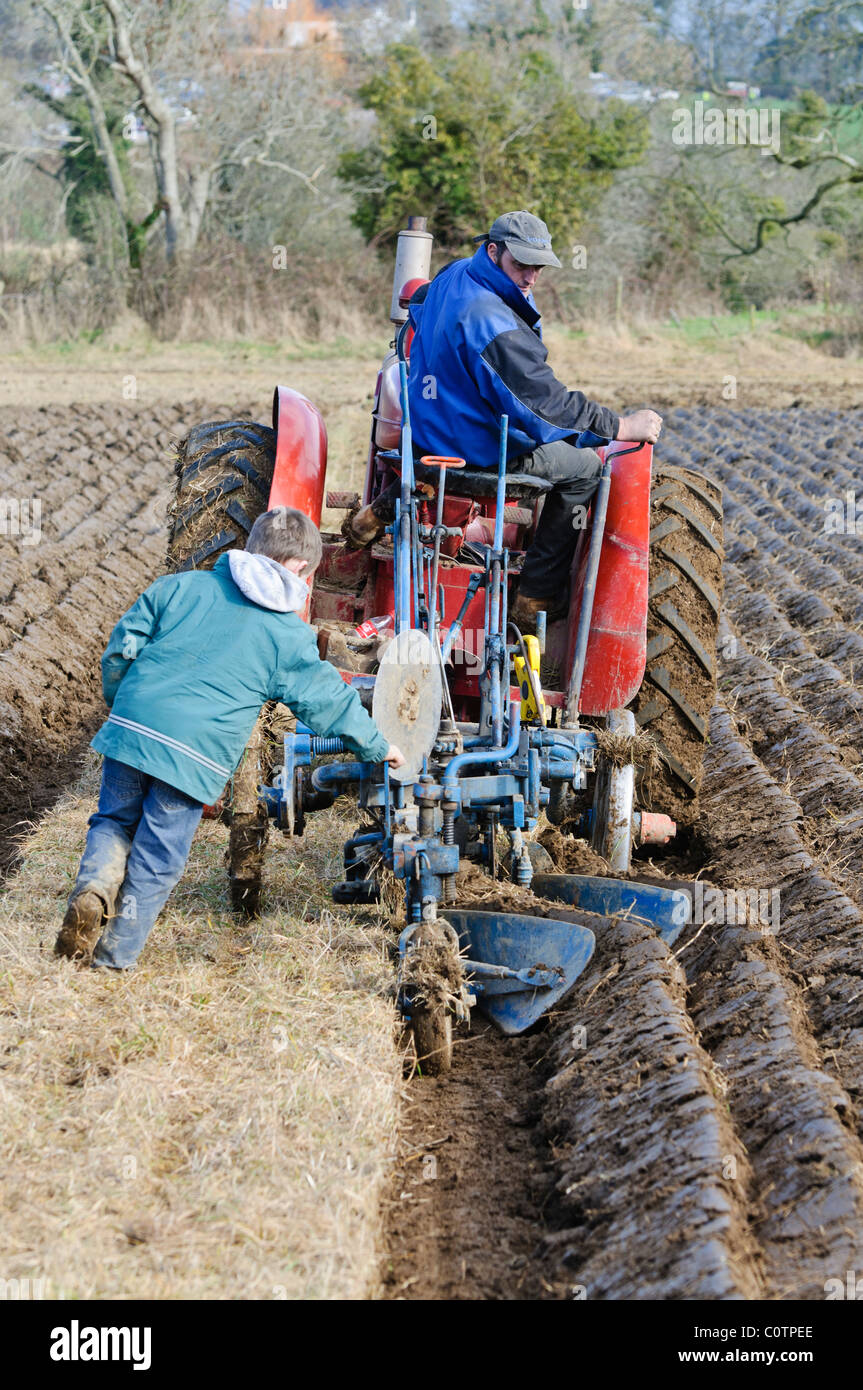 L'agricoltore utilizza un trattore d'epoca per arare un campo mentre suo figlio assiste Foto Stock