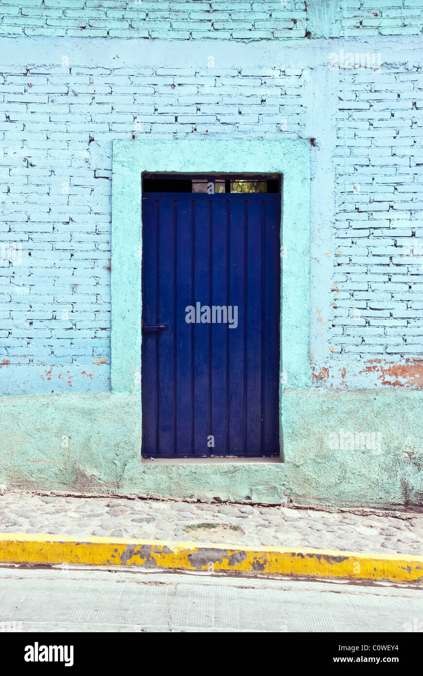 Il cerulean blu dipinto di un muro di mattoni con Deep Blue Door marciapiede di pietra e di colore giallo brillante cordolo Teotitlan marcatura dello Stato di Oaxaca Messico Foto Stock