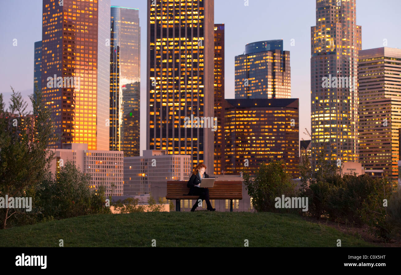 Donna seduta su una panchina nel parco lavorando sul computer portatile con Los Angeles skyline della città sullo sfondo al tramonto tramonto Foto Stock