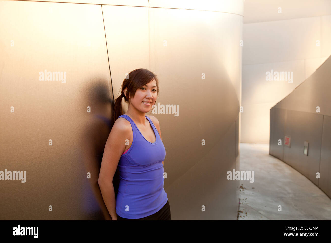 Americano asiatico femminile in appoggio appoggiata contro metal clad parete. Viola tank top e pantaloni neri. Foto Stock