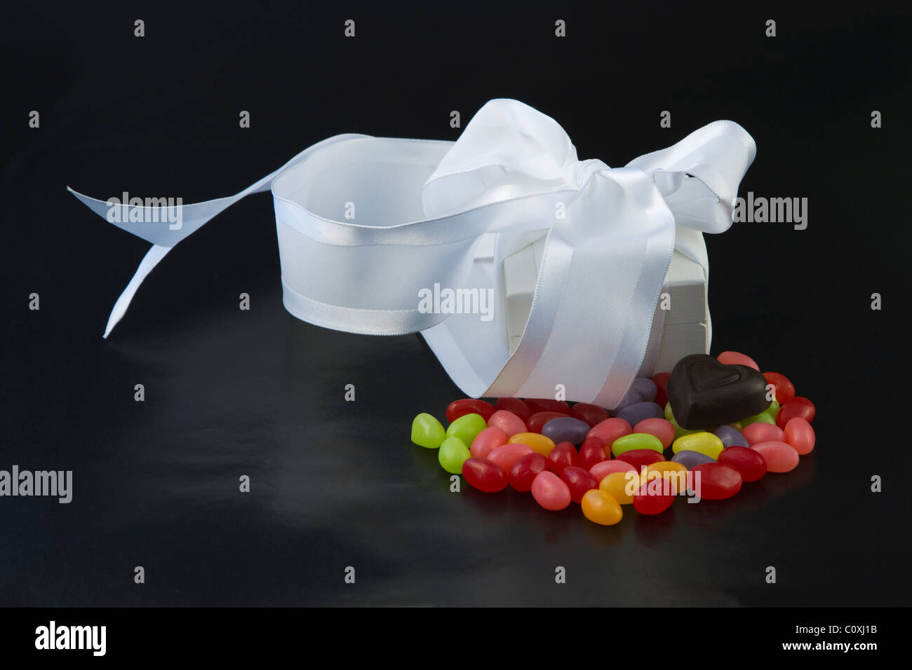 White box gioielli avvolto con il bianco di raso collocato sulla colorata jellybeans con cuore di cioccolato su sfondo nero. Foto Stock