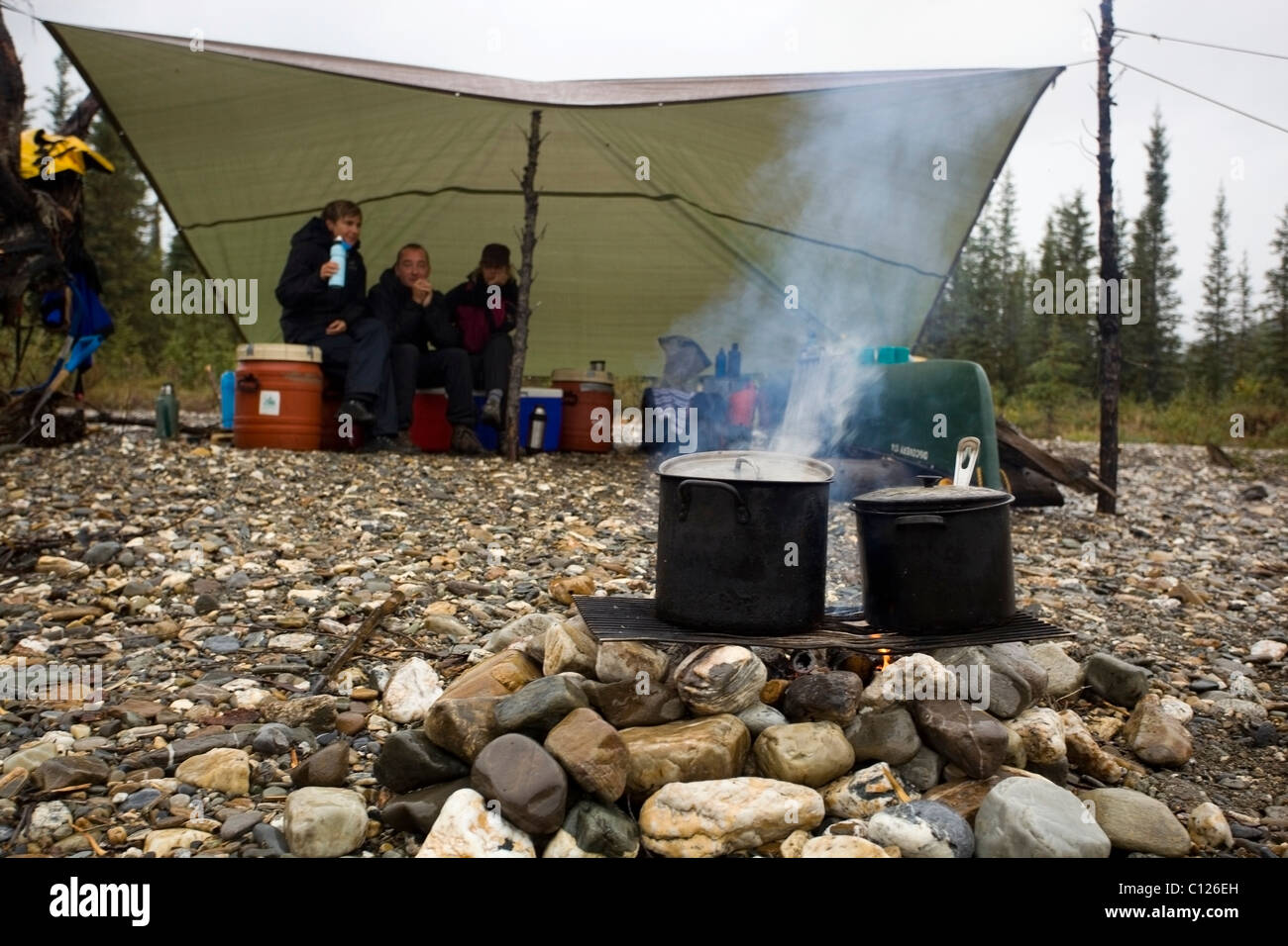 La cottura su un fuoco di campo, falò, grill, pentole, vapore, gente seduta sotto tarp, pioggia, superiore Liard River, Yukon Territory, Kanada Foto Stock
