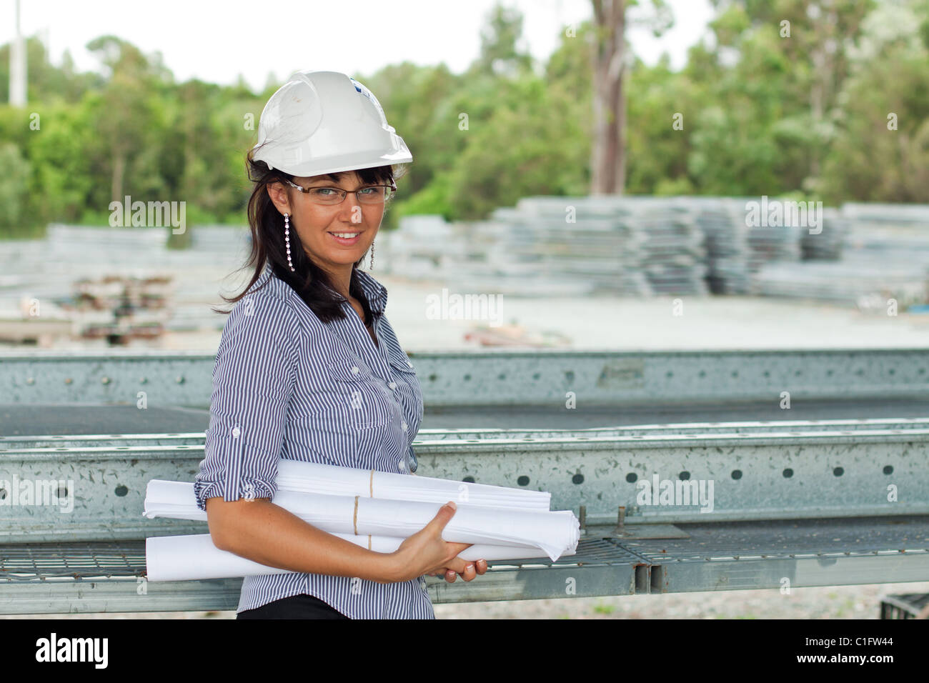 Sorridente giovane ingegnere donna con disegni arrotolati nelle mani di chi indossa occhiali e casco in magazzino Foto Stock