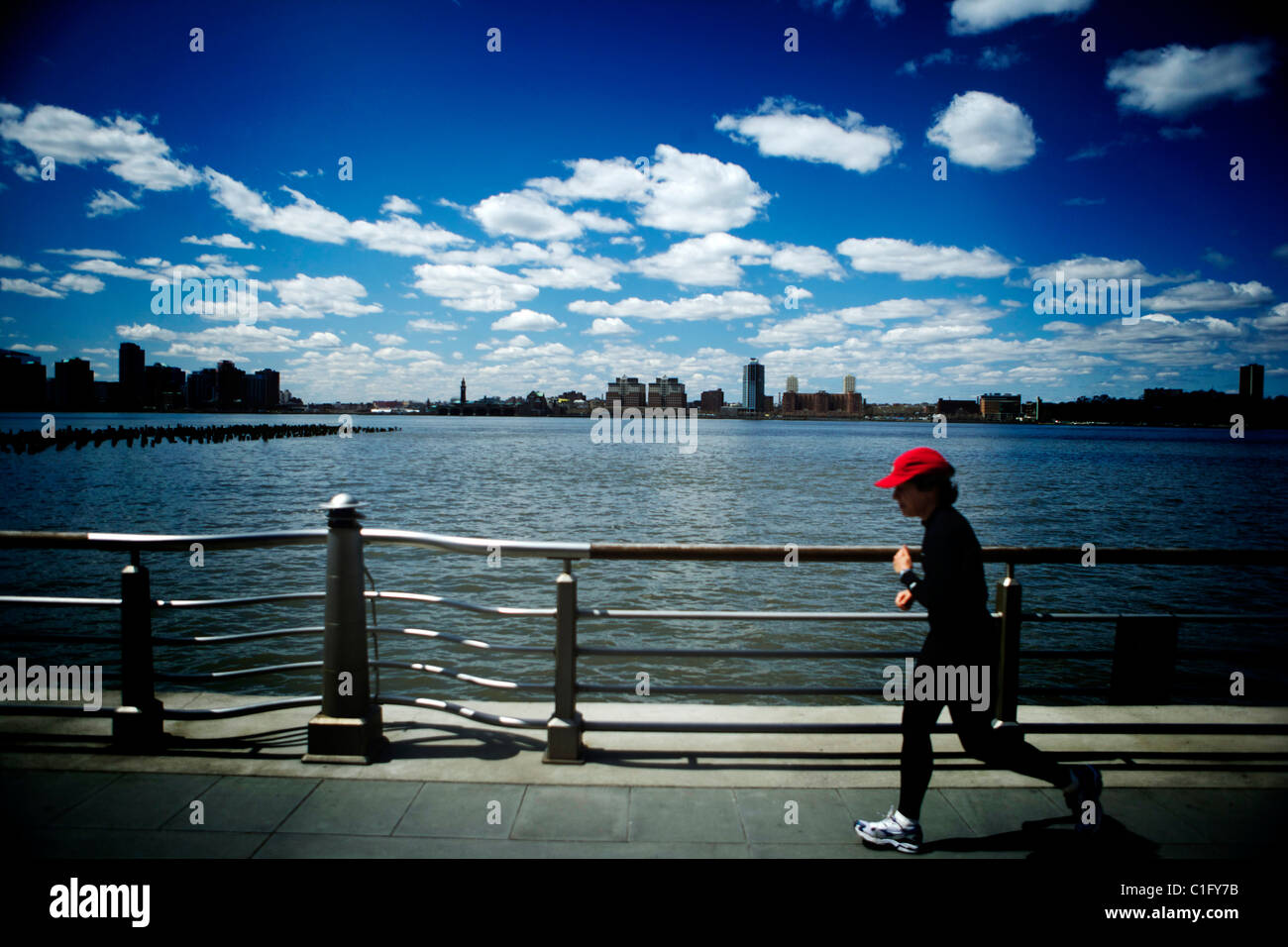 Un pareggiatore di runner in un rosso berretto da baseball con lo skyline di New York in background in esecuzione lungo il fiume Hudson. Foto Stock