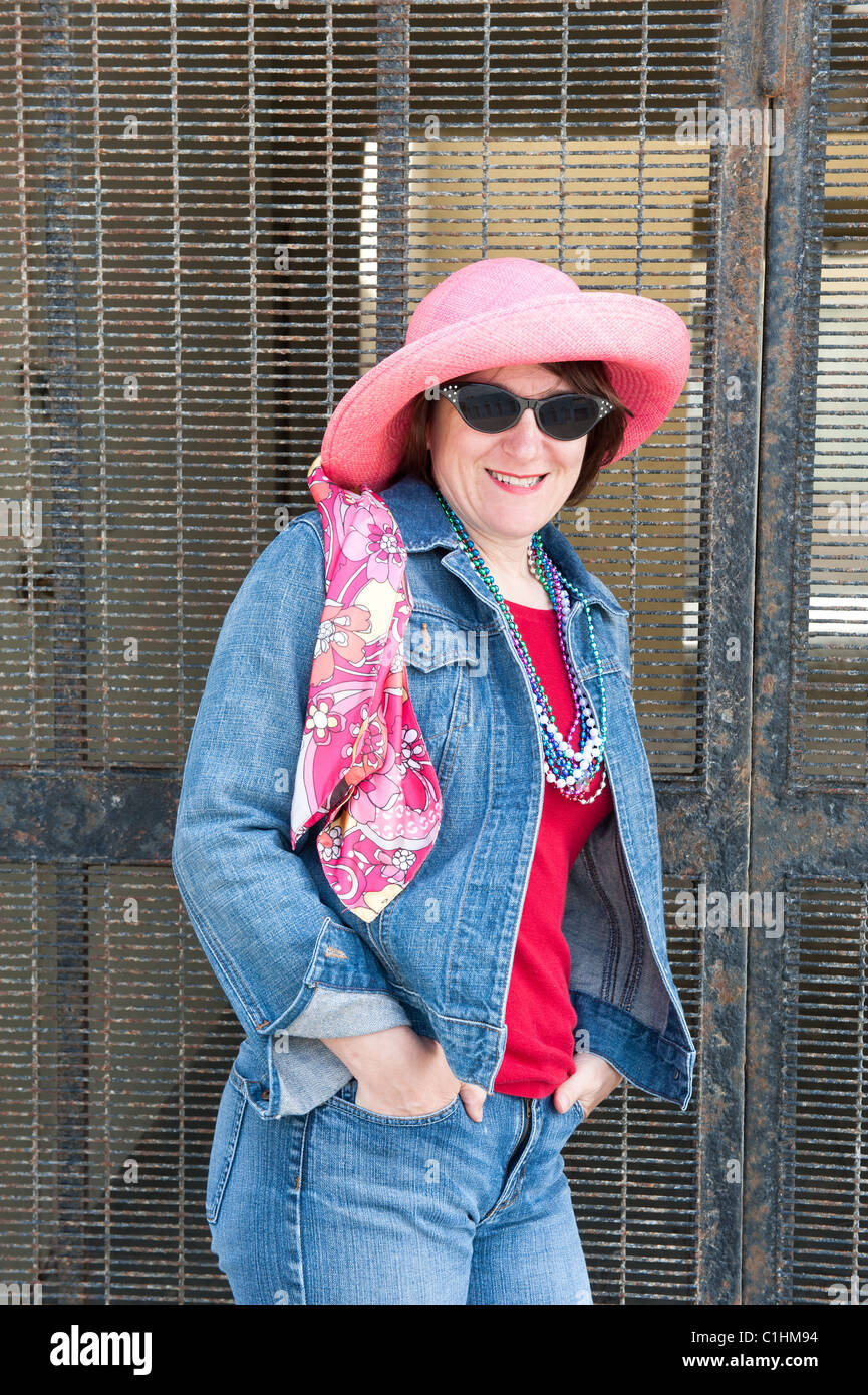 Una donna pone contro un metallo cancello di sicurezza mentre si indossa un colorato cappello rosa vibranti, sciarpa e occhiali da sole. Foto Stock