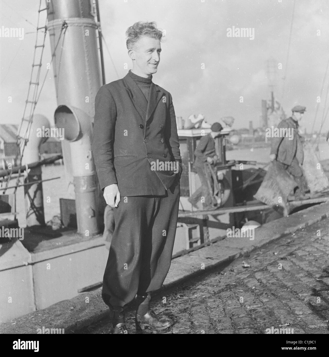 1950, un docker maschio in piedi fuori su una banchina acciottolata, Newcastle-upon-Tyne, Inghilterra, altri portuali che tengono sacchi che attraversano su una tavola da una barca. Foto Stock